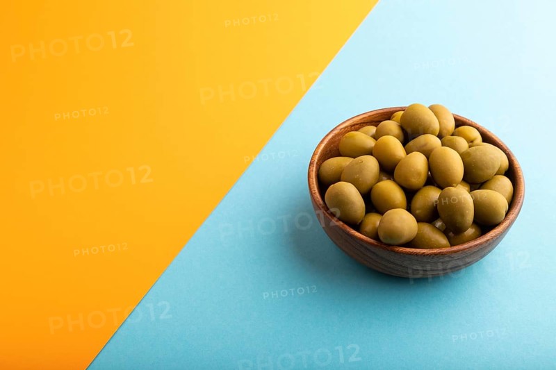 Fresh green olives in wooden bowl on blue and orange pastel paper background. side view, copy space