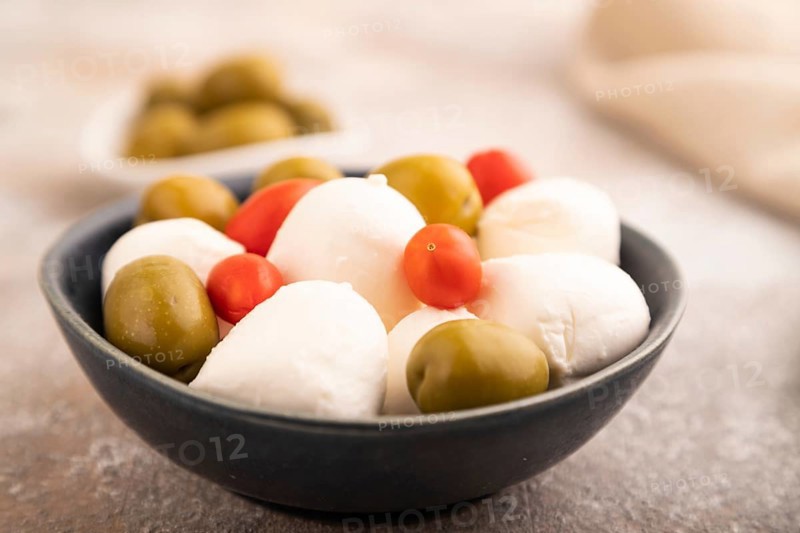 White Mozzarella cheese, with tomatoes and olives in blue bowl on brown concrete background and linen textile, side view, close up, selective focus