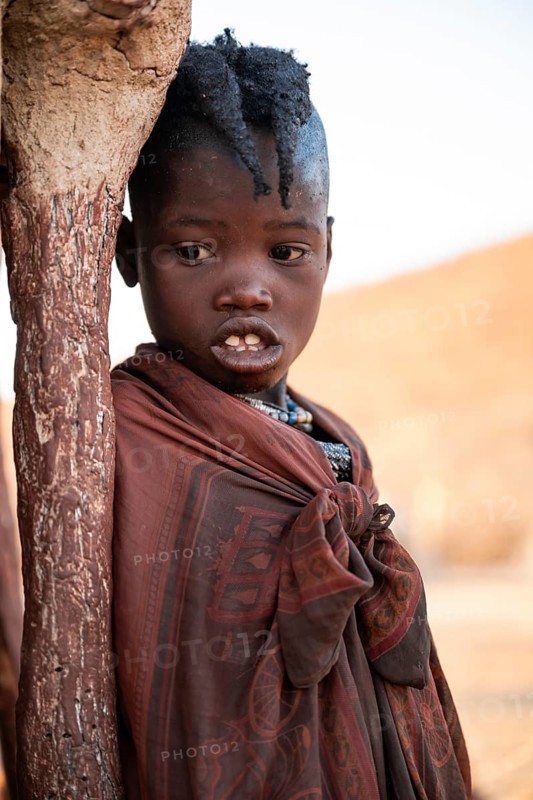 Himba girl leaning at a traditional clay hut, in the morning, traditional Himba village, Kaokoveld, Kunene, Namibia