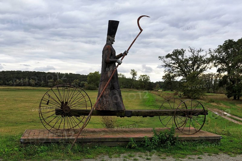 Sculpture in a meadow, traditional grim reaper, figure carrying a scythe, standing on cart with large wheels, symbol of death, sculpture of ferryman Charon by Bernd Streiter on the Elbe cycle path, twilight, Mödlich, Prignitz, Brandenburg, Germany