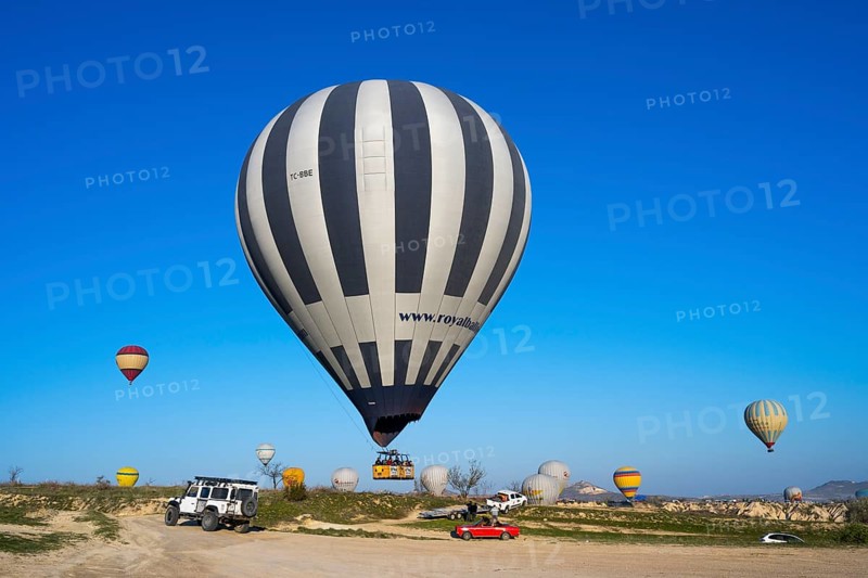 Large striped hot air balloon lands next to jeep and red car in clear landscape, Göreme Tarihî Millî Park, Nevsehir Province, Nevsehir, Cappadocia, Capadocia, Cappadocia, Central Anatolia, Turkey