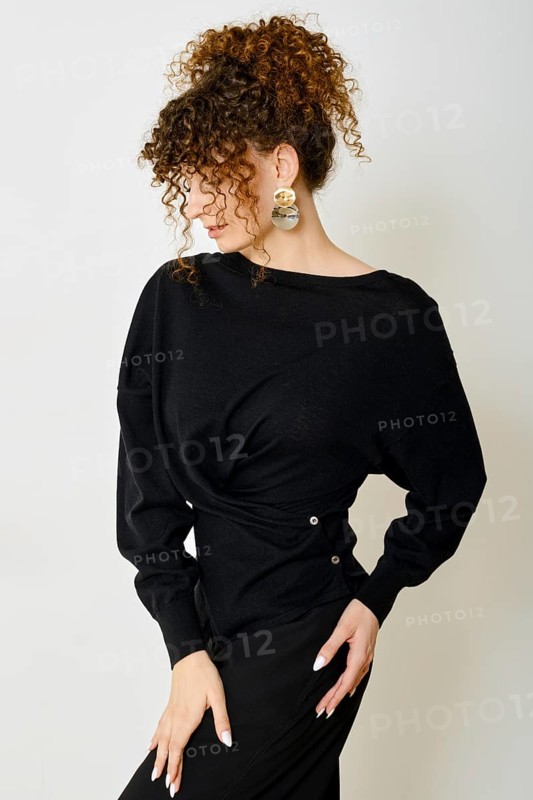 A stylish model with curly hair poses against a neutral backdrop. She wears black blouse with twist and accentuates her look with bold accessories. The overall vibe is chic and modern