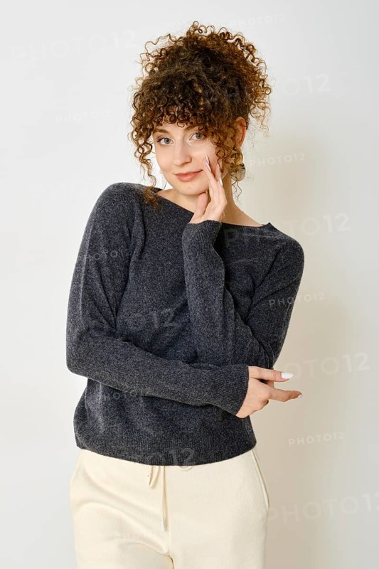 Cheerful woman in dark sweater and light pants standing against white wall with a playful expression