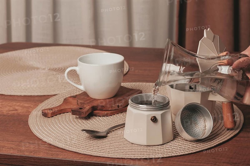 Hot water is being poured from a glass kettle into a coffee maker on a wooden table. A white cup awaits the fresh brew in a simple kitchen environment