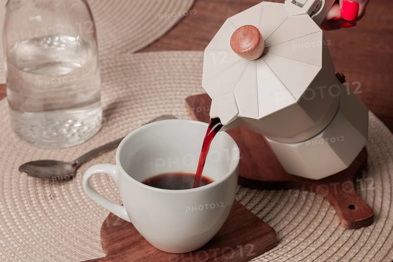 Freshly brewed coffee is being poured from a stovetop espresso maker into a white cup on a wooden table. A clear glass water bottle sits nearby, adding to the warm, inviting setting