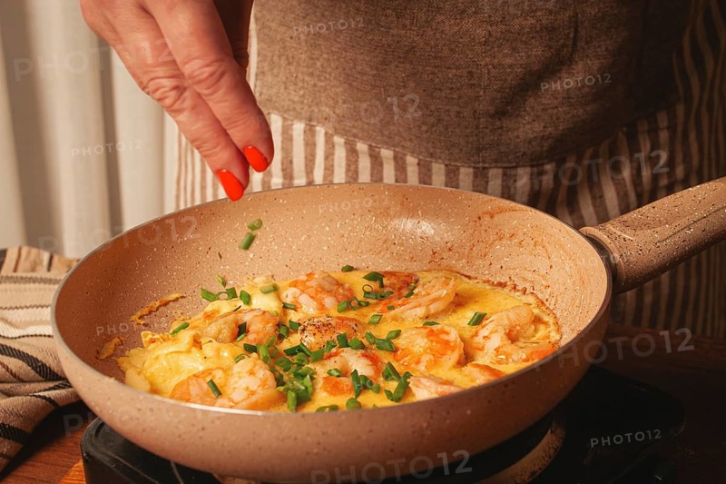 A woman is cooking shrimp in a skillet, adding fresh herbs to the dish. The warm colors of the kitchen create a cozy atmosphere while she prepares dinner