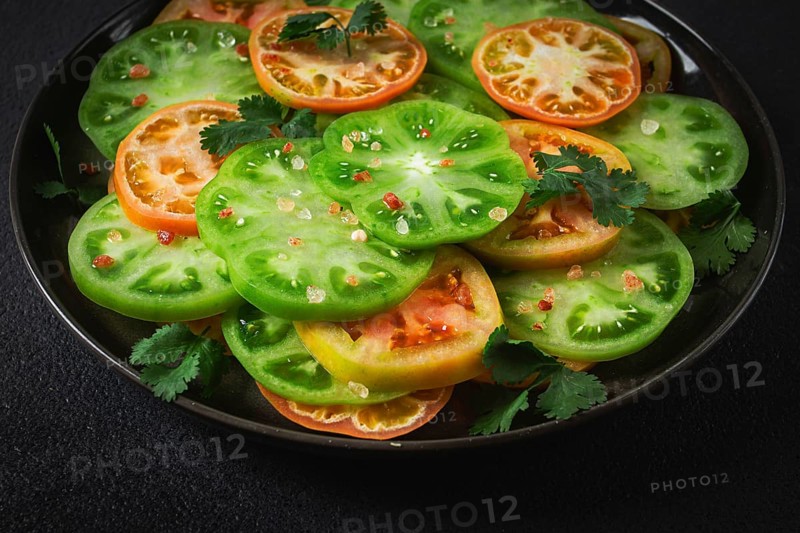 Sliced green tomatoes, on a dark surface, garnished with herbs and salt, top view