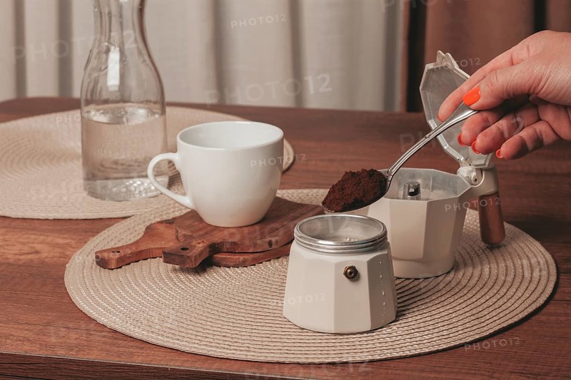 A hand holds a spoonful of coffee grounds over a moka pot while a cup and a bottle of water sit on a wooden table. The scene is cozy and inviting, ideal for coffee lovers