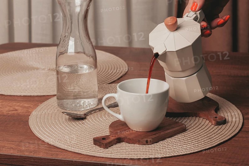 A hand pours freshly brewed coffee from a stovetop maker into a white cup. A glass pitcher filled with water is nearby on a wooden table with natural textures