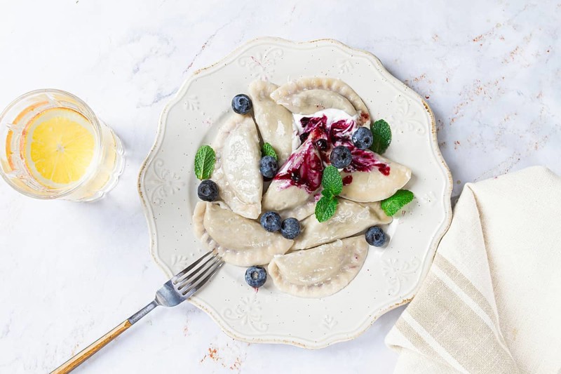 Handmade dumplings filled with sweet ingredients, arranged beautifully on a white plate, topped with berry sauce, fresh blueberries, and mint leaves, ready to enjoy