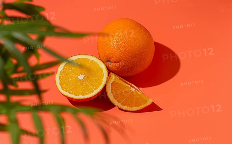 On the coral surface, next to two neatly sliced slices, there is a whole orange. Sunlight casts soft shadows, emphasizing the freshness and bright color of the fruit