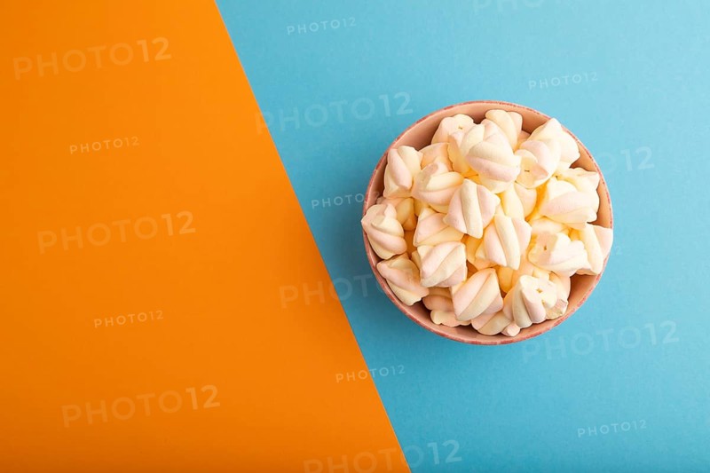 Orange and pink marshmallow in ceramic bowl on blue and orange pastel paper background, top view, flat lay, copy space, minimalism
