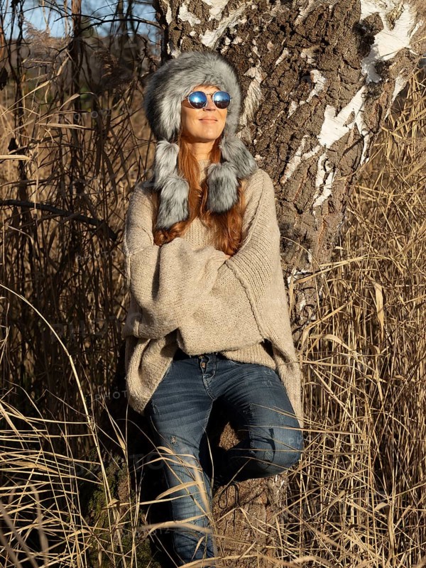 Slim woman with fur hat and sunglasses, red hair and warm winter colors between birch forest and reeds