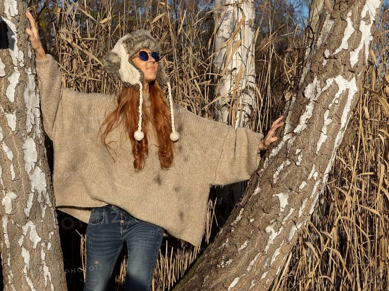 Slender pretty woman with fur hat and sweater, red hair and warm winter colors between birch forest and reeds
