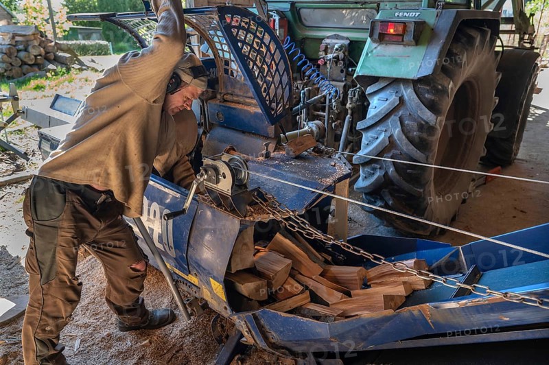 Young man sawing and splitting beech logs with his saw cutting machine, powered by a tractor in a barn, Othenstorf, Meckleburg-Vorpommern, Germany