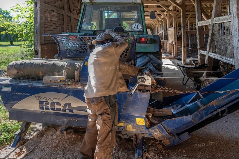 Young man sawing and splitting a beech log with his saw cutting machine with a conveyor belt, driven by a tractor in a barn, Othenstorf, Meckleburg-Vorpommern, Germany