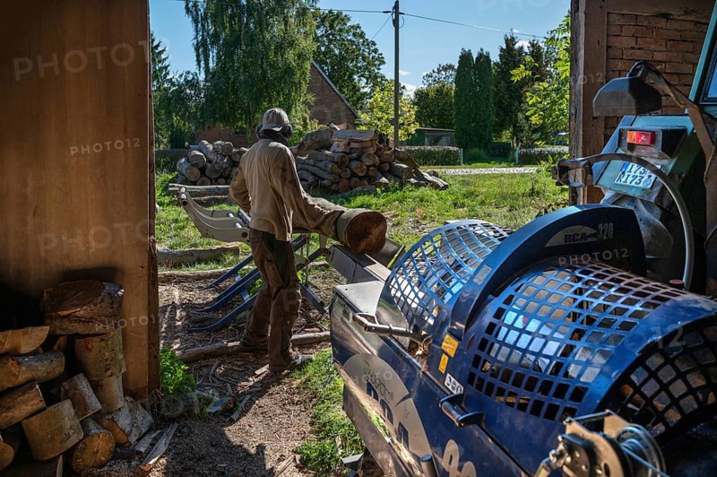 Young man sawing and splitting beech logs with his saw cutting machine, powered by a tractor in a barn, Othenstorf, Meckleburg-Vorpommern, Germany