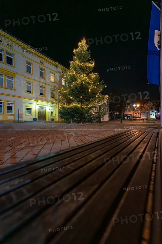 A brightly lit Christmas tree next to a building and a wooden bench, Aidlingen, Böblingen district, Germany