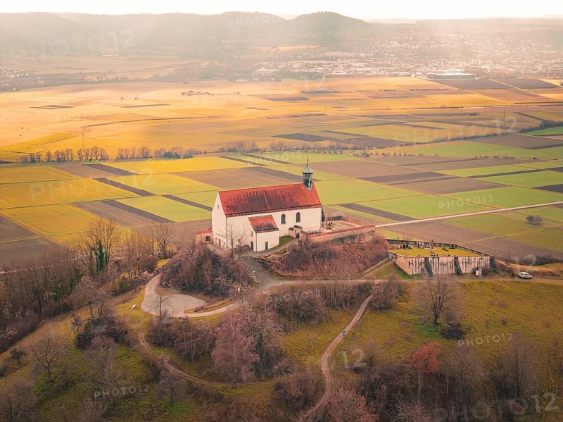 A church with a red roof towers over a vast mosaic of fields in sunlight, Wurmlingen Chapel, Rottenburg am Neckar, Germany