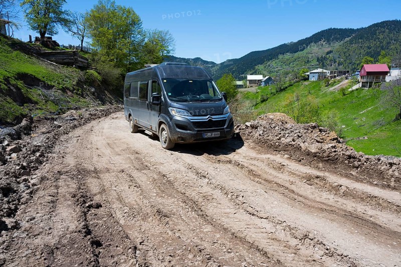 A van drives down a muddy road surrounded by green hills and individual houses, campers on the way to Goderdzi Pass, Ajara region, Adjara, Autonomous Republic, Georgia