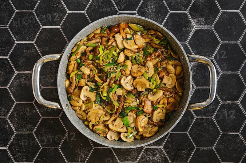 Freshly cooked mushrooms and onions are sautÃ©ed in a large skillet. Green leek add flavor. The meal is prepared in a stylish kitchen with a hexagon tile backdrop during lunch time