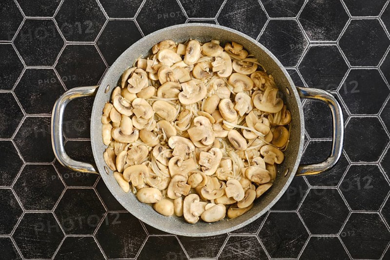 Sliced mushrooms are being fried in a large pan, giving off a savoury aroma. The scene is set on a stylish black hexagonal countertop
