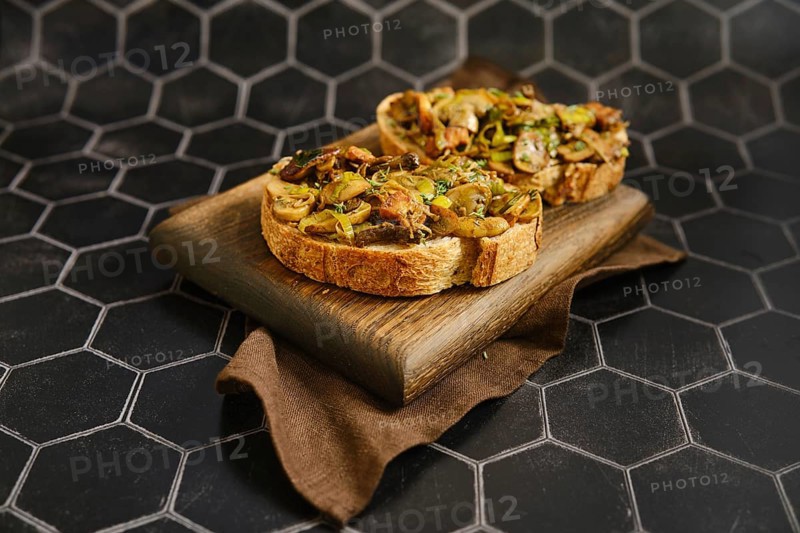 Freshly made mushroom sandwich features honey mushroom, enoki mushroom, champignon, and leek on crusty bread. Served on a wooden board, it looks inviting against a dark patterned background
