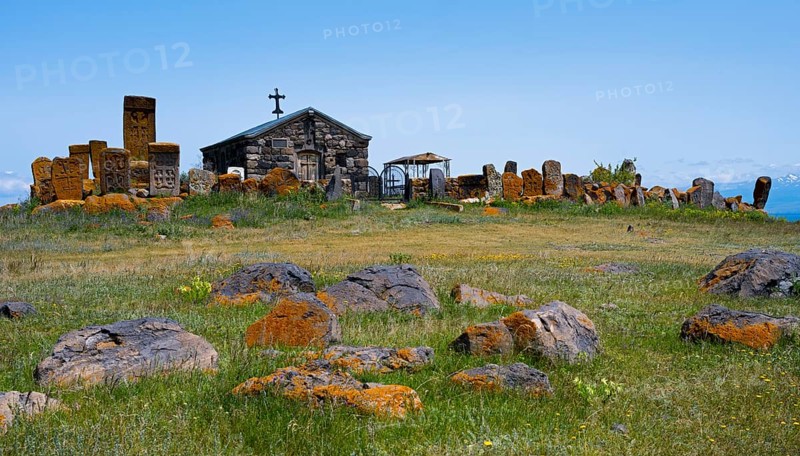 Small chapel surrounded by old tombstones on a hilly meadow under a blue sky, khachkar at the chapel of the Holy Cross and cemetery near Noratus, Sarukhan, Lake Sevan, Gegharkunik province, Armenia