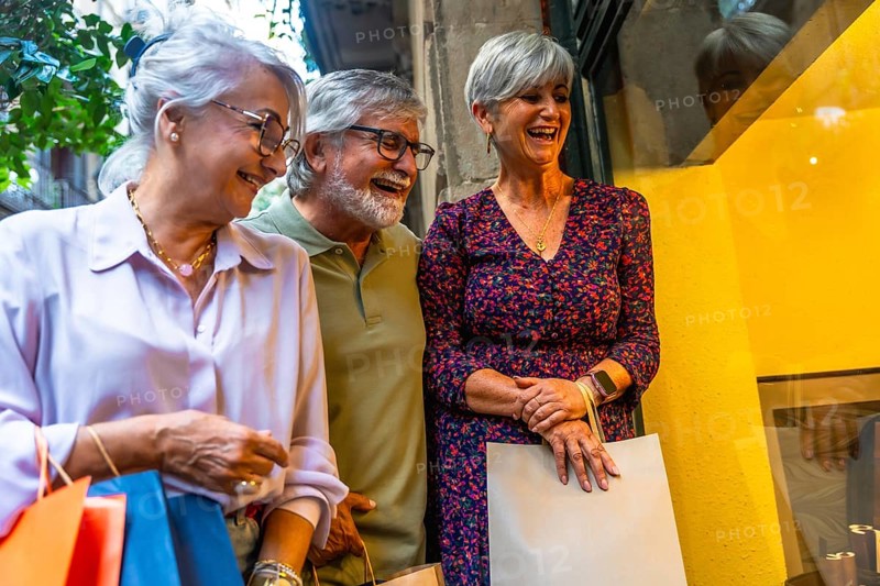Senior friends laughing together while window shopping on a city street, carrying multiple shopping bags and enjoying their leisure time during retirement