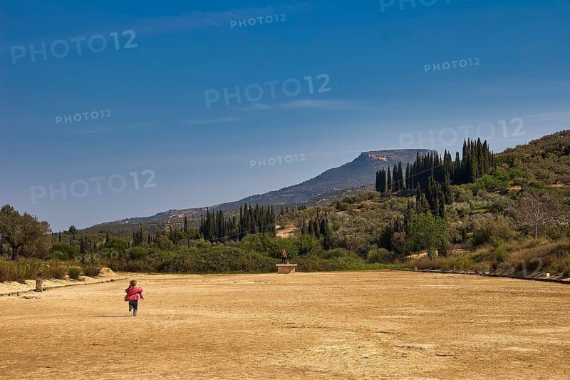 A child runs across a dry meadow to distant mountains, under a clear sky with cypresses in the background, Ancient Stadium, Nemea, Peloponnese, Greece