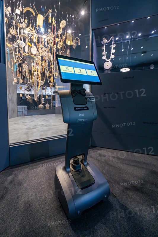 A robot in front of a glass display case full of jewelry in a dark exhibition area, robotic personality Roboloutions in the jewelry museum, Pforzheim, Germany