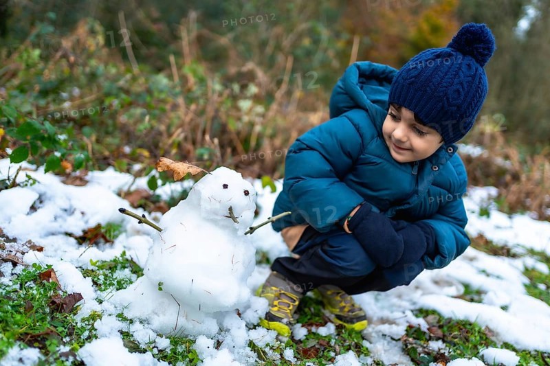 Young boy kneeling on snowy ground, wearing a warm coat, hat, and gloves, observing a freshly made small snowman with a happy expression during a cold winter day