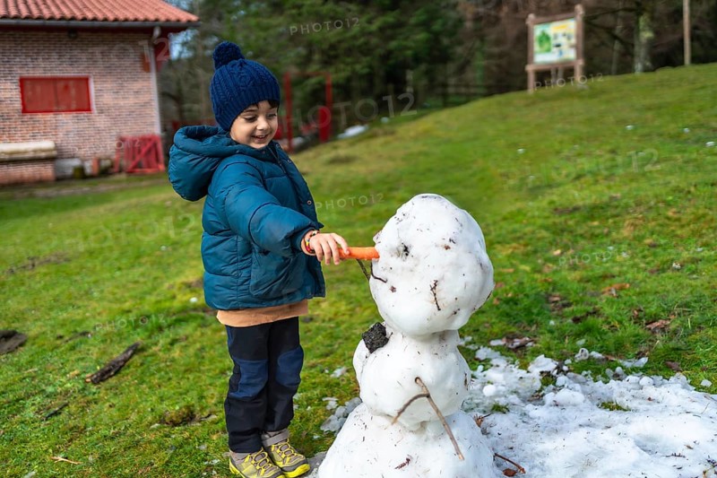 Young boy smiling and decorating a melting snowman with a carrot nose, wearing a warm hat and jacket while playing in a snowy patch on a green grassy hill
