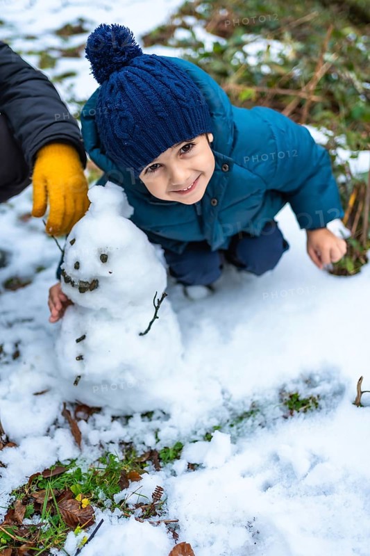 Young child wearing a blue knitted hat and warm winter clothes kneeling in the snow, proudly interacting with a small snowman built outdoors on a cold crisp day