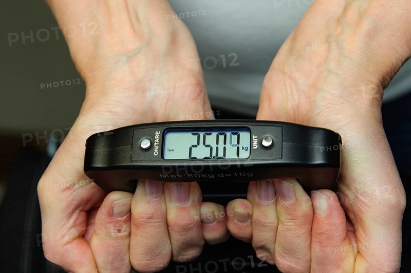 Woman holding hand scale for weight of suitcase travel bag flight luggage checks luggage weight for excess luggage in front of flight travel, scale shows 25.09 kg kilos kilograms, international