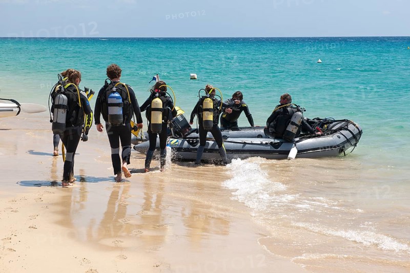 Recreational divers walk across beach to inflatable boat in diving suits with scuba tanks on their backs for boat trip trip to dive site off the coast, East Atlantic, Jandia Peninsula, Fuerteventura, Canary Islands, Spain