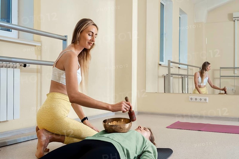 A yoga instructor assists a student in a serene studio setting, using a singing bowl during a relaxing session