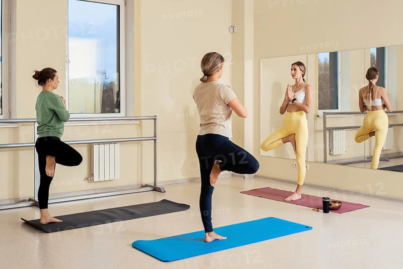 Three women practice yoga in a well-lit studio, showcasing various poses that emphasize balance. A large mirror reflects their focus and determination during the session
