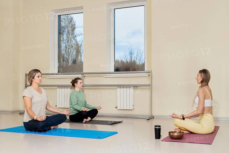 Three women are seated on yoga mats in a bright studio. They are focusing on meditation, with one instructor guiding them. The serene atmosphere promotes mindfulness and relaxation