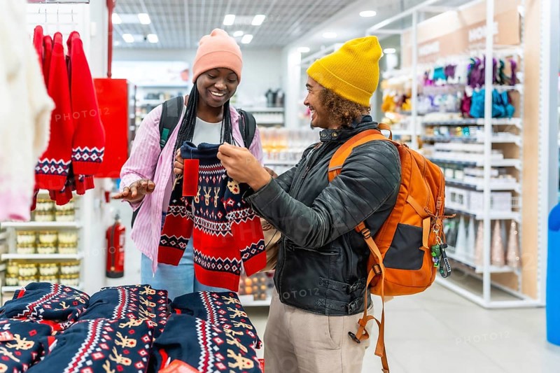 Happy young multiracial couple smiling as they browse and select red holiday sweaters in a busy retail store, enjoying festive shopping, bonding and seasonal gift buying