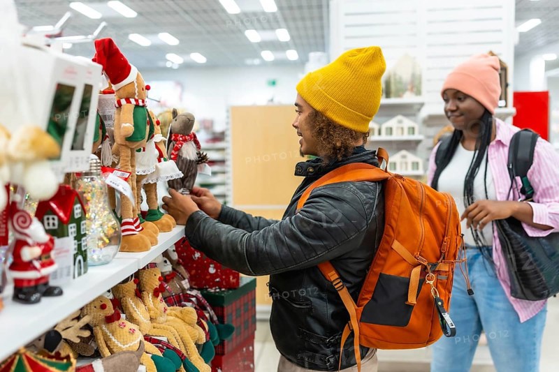 Young man and woman choosing holiday gifts and ornaments, enjoying the festive atmosphere while looking at various seasonal items on display in a modern shopping mall