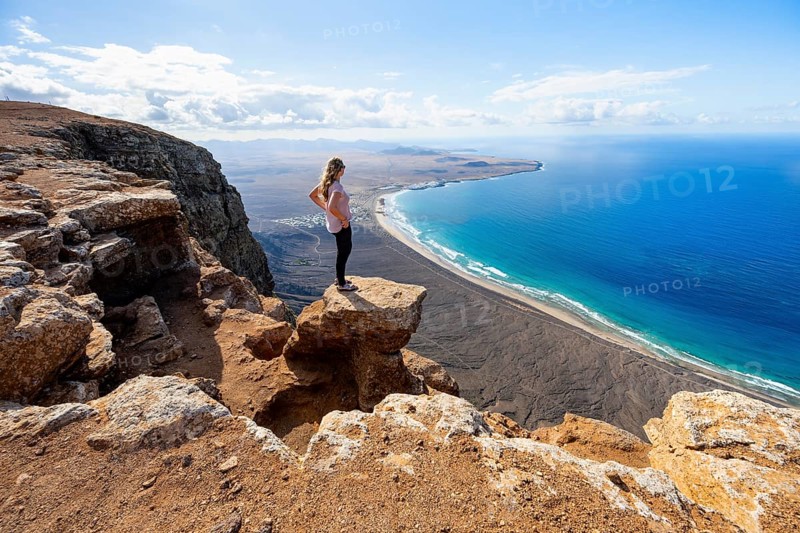 Young woman enjoying the view from the Risco de Famara cliffs to Famara beach, Playa de Famara with La Calaeta, Lanzarote, Canary Islands, Spain