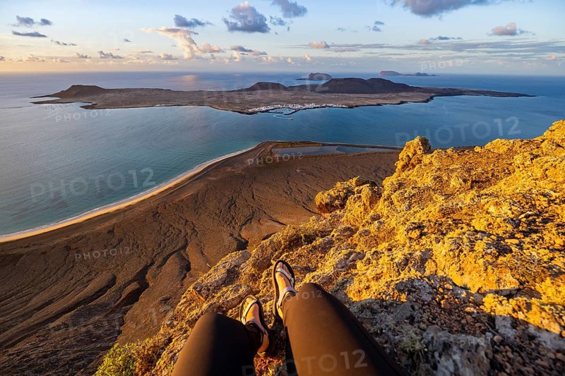 First person view, legs of a woman sitting on a steep cliff, view of the sea and the island of La Graciosa, Mirador del Porrito viewpoint at sunset, Lanzarote, Canary Islands, Spain