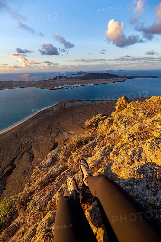 First person view, legs of a woman sitting on a steep cliff, view of the sea and the island of La Graciosa, Mirador del Porrito viewpoint at sunset, Lanzarote, Canary Islands, Spain