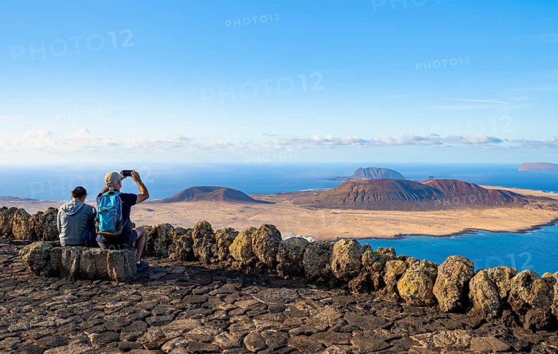 Tourists on a bench on the viewing platform at the Mirador del RÃ­o viewpoint, in the evening light, view of the sea and of the island of La Graciosa with volcanic craters, designed by artist César Manrique, Lanzarote, Canary Islands, Spain
