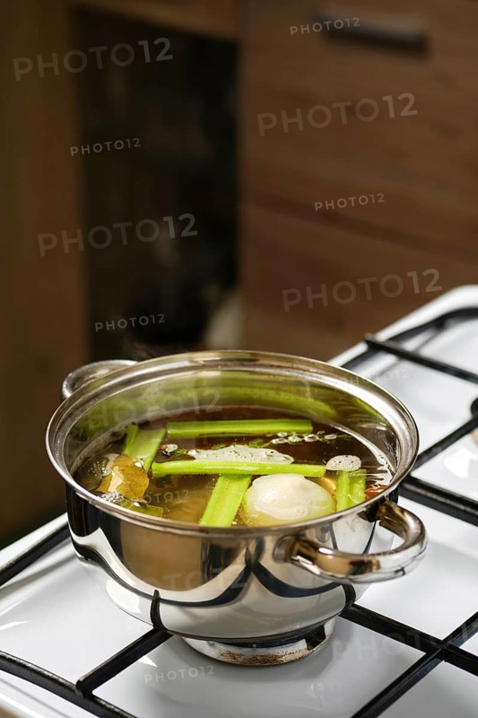 Cooking vegetable broth on the stove in a stainless steel pot with fresh celery, onion, carrot, bay leaf and peppercorns. Steam rises from the pot, indicating a warm cooking environment during meal preparation