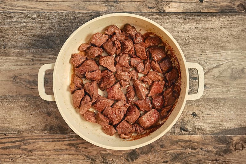 Chunks of beef are searing in a large pot on a weathered wooden table. The meat is cooked to a rich brown color, suggesting a flavorful dish is being prepared