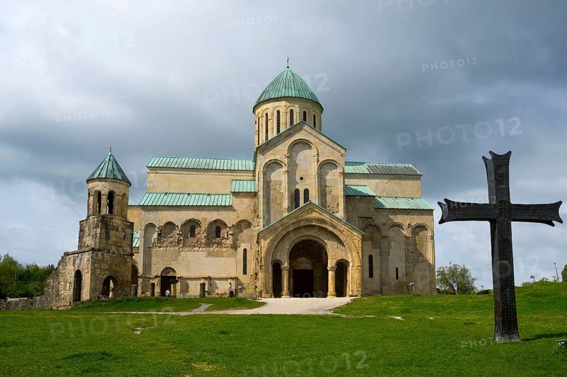 Historic church under a cloudy sky surrounded by greenery with a large cross in the foreground, Bagrati Cathedral, Church of the Dormition of the Most Holy Mother of God, UNESCO World Heritage Site, Kutaisi, Imereti Region, Georgia