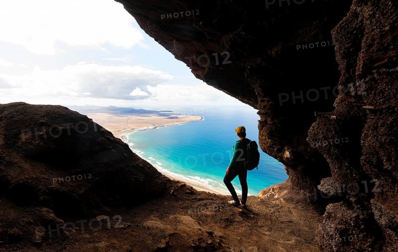 Tourist in the Cueva de las Cabras cave, young woman enjoying the view from the Risco de Famara cliffs on Famara beach, Playa de Famara with La Calaeta, Lanzarote, Canary Islands, Spain