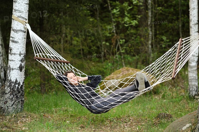 A woman enjoys a peaceful afternoon in a hammock, nestled between trees. She has a relaxed posture, with the serene green landscape creating a calming atmosphere in the woods